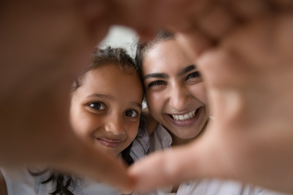 A woman and a young girl smiling with their hands forming a heart in front of the camera lense 