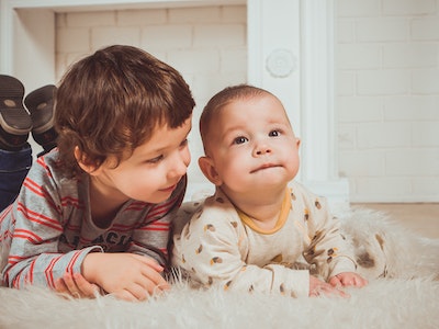 A baby lying on a rug next to a toddler