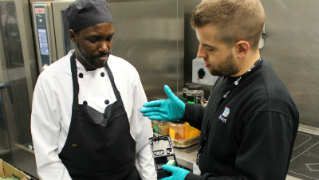 Two men in a kitchen, one is wearing an apron and a hat and one is talking to the other man